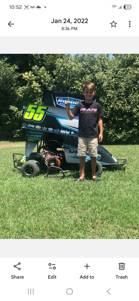 Young boy standing with a blue and black number 55 sprint racing car on grass with green hedges in background
