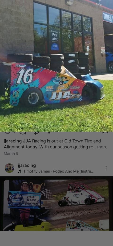 Blue and red race car number 16 parked on grass in front of tire shop building