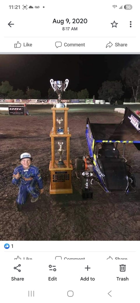 Young child in blue racing suit standing next to large golden trophy at night dirt track racing event
