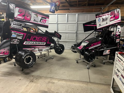 Two black sprint cars with pink accents displayed in a garage, equipped with large tires and racing components.