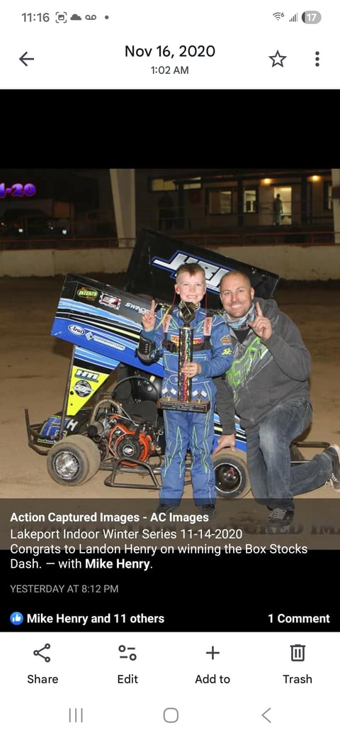 Young race car driver and adult man pose with trophy next to blue sprint car at indoor racing venue